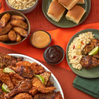 A closeup spread of Pollo Campero's menu items on an orange table, including rice, buns, roasted potatoes, sauce, grilled chicken, and fried chicken.