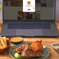 A closeup of a desk with a laptop open and the screen displaying the Pollo Campero order online window. In front of the laptop is a Pollo Campero grilled and fried chicken meal with sauce and fries.