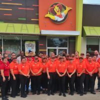 A wide photograph of Pollo Campero chicken franchise staff, wearing red polo shirts, black pants, and brown hats, standing outside of a Pollo Campero location.